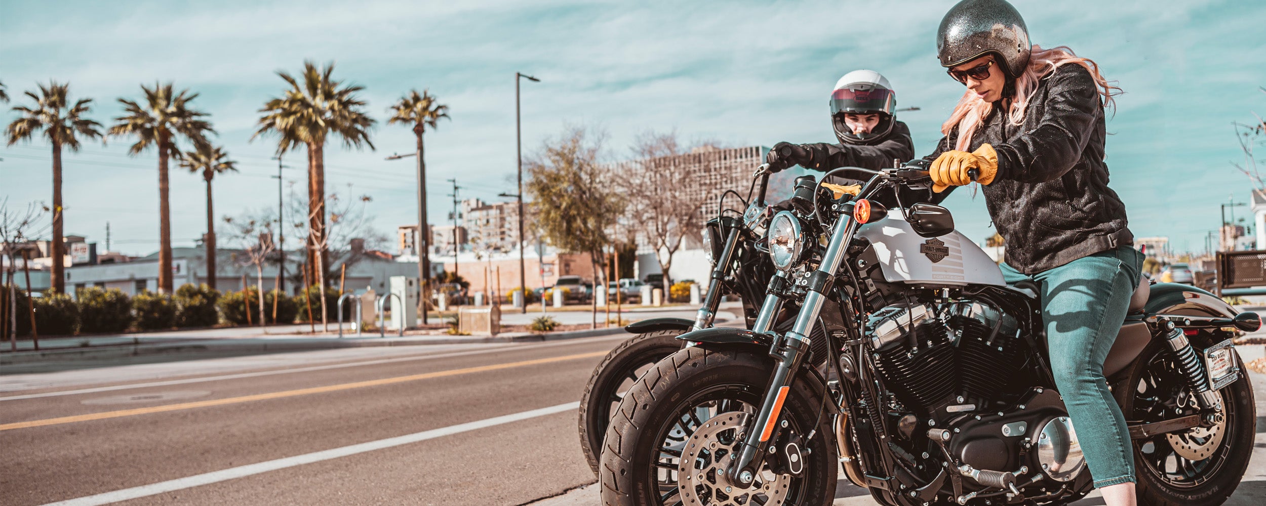 Two people with a motorcycle on a city street with palm trees in the background