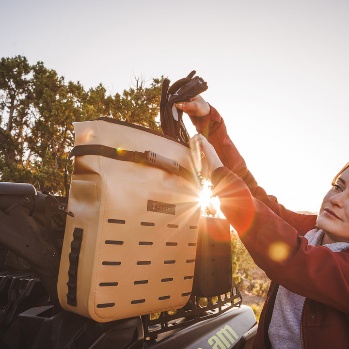 Person attaching a beige Molle Bag to a UTV with trees and sunlight in the background