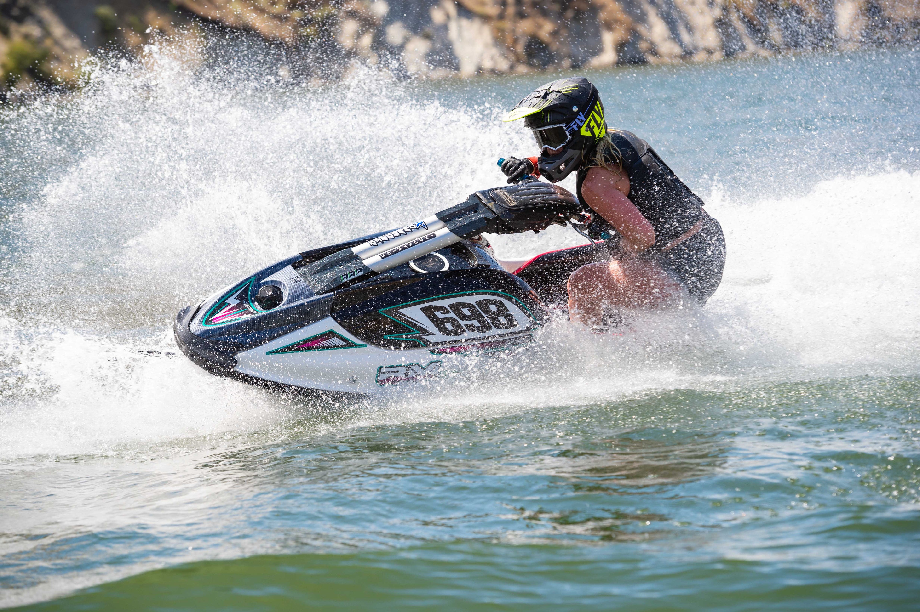 A person riding a black, white, and teal jet ski across choppy water, creating a large spray of white foam. The rider is wearing a black helmet and life vest, and the background shows a rocky shoreline.