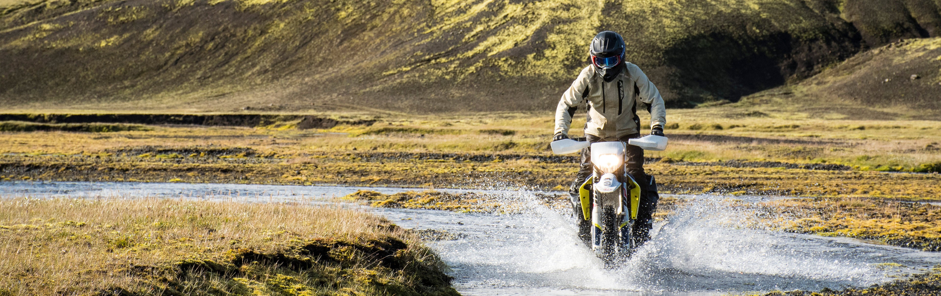 Person riding a motorcycle through a shallow stream with mountains in the background