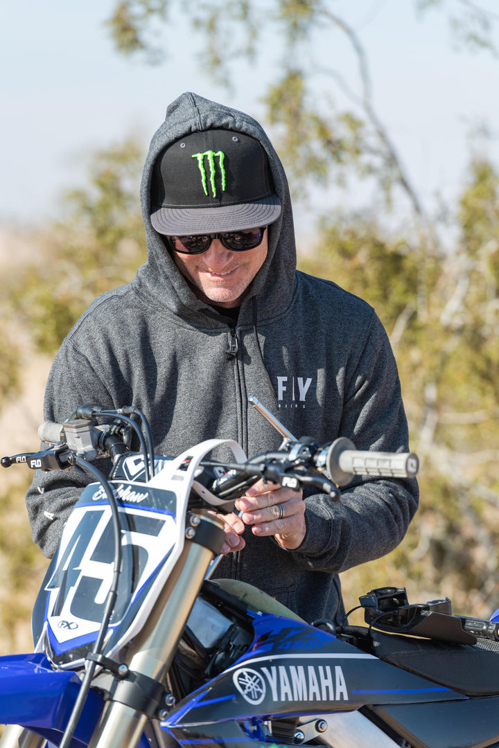 Person wearing a hoodie with a Monster Energy logo, sitting on a Yamaha dirt bike outdoors.