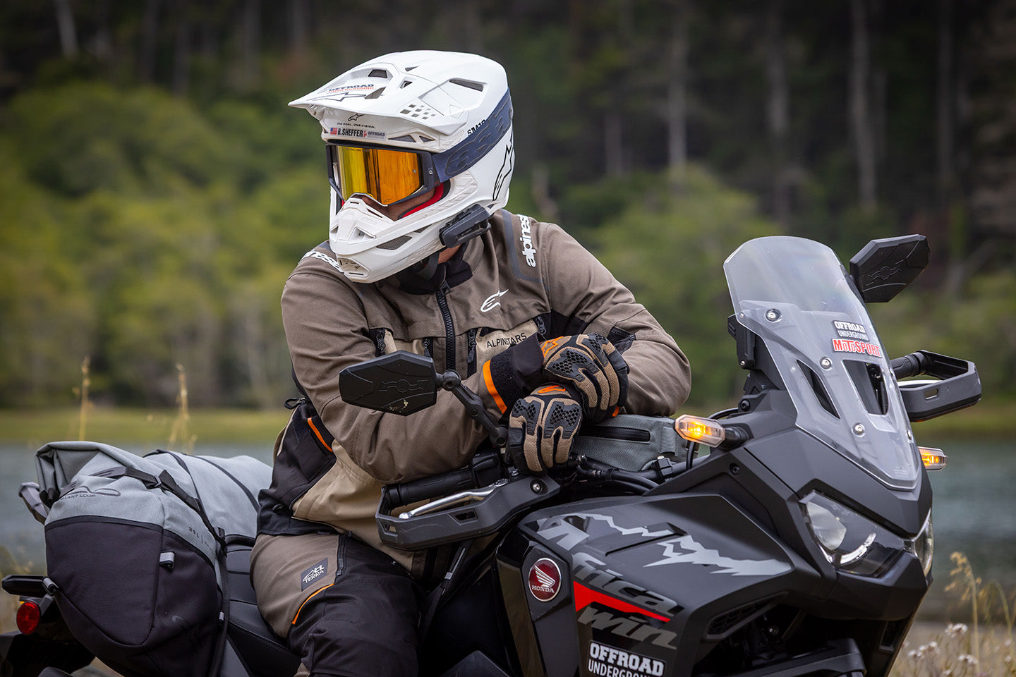 A person wearing a white full-face helmet with orange-tinted goggles, a brown riding jacket, and gloves sits on a black off-road motorcycle outdoors near a body of water with a forest in the background.