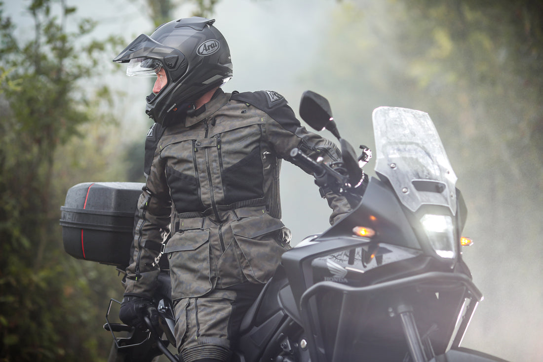 Person in motorcycle gear sitting on a motorcycle with a helmet on, surrounded by trees.