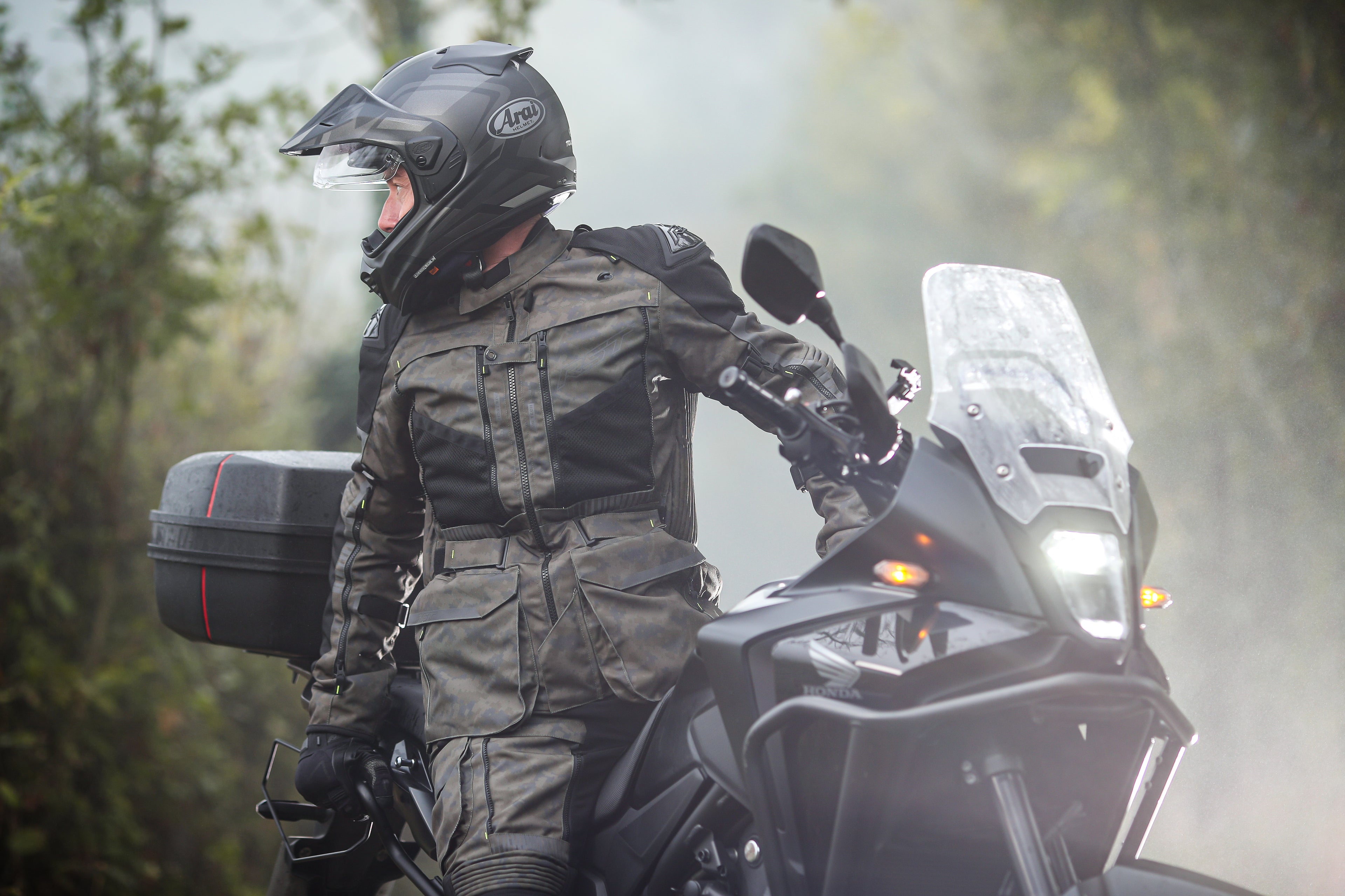 Person in motorcycle gear sitting on a motorcycle with a helmet on, surrounded by trees.